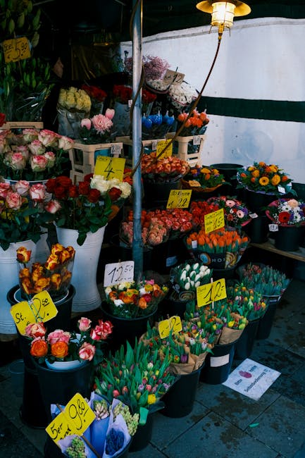 A vibrant display of fresh flowers at an outdoor stall, arranged in black, white, and clear containers. The selection includes pink roses with lush green stems, bright orange and yellow sunflowers, an