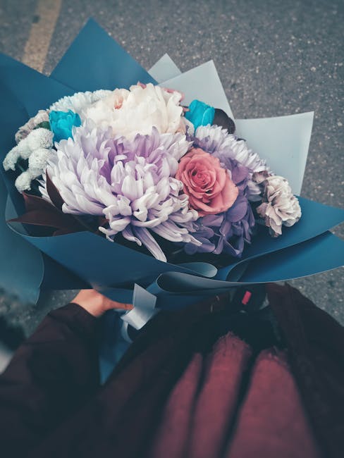 A hand holding a bouquet of fresh flowers wrapped in matte dark blue paper, featuring a large white and light purple chrysanthemum at the center, accompanied by a pink rose, violet irises, and smaller