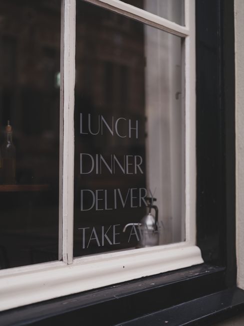 A close-up of a white-painted window frame with glass panes, displaying a reflection of an interior space. On the glass, there are white printed words listing services: 'LUNCH,' 'DINNER,' 'DELIVERY,' 