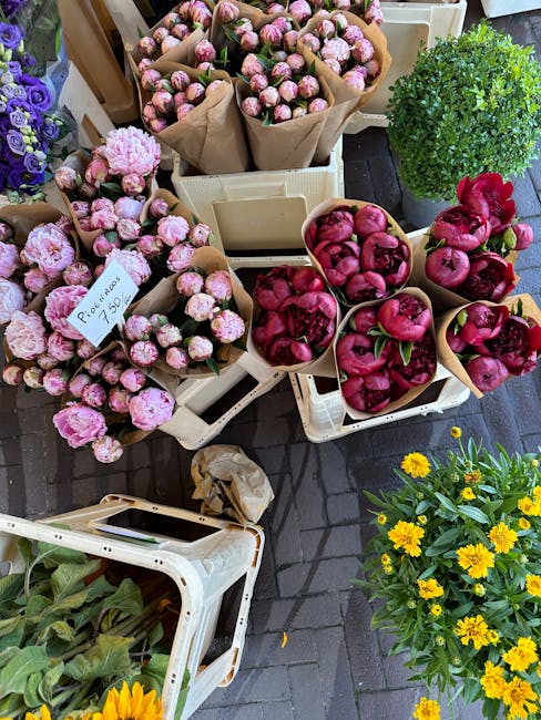 A display of fresh flower bouquets and potted plants arranged on white plastic crates outside a florist shop. The bouquets consist of pink peony flowers wrapped in brown paper, with some in containers