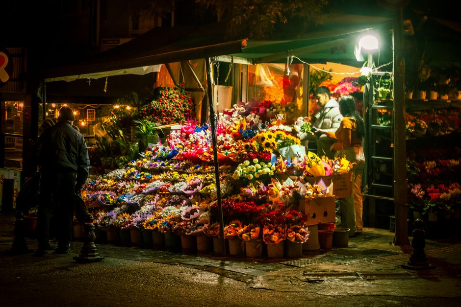 A vibrant outdoor flower market at night featuring a wide variety of fresh flowers arranged in buckets and bouquets, including sunflowers, roses, lilies, and chrysanthemums in bright colors such as ye