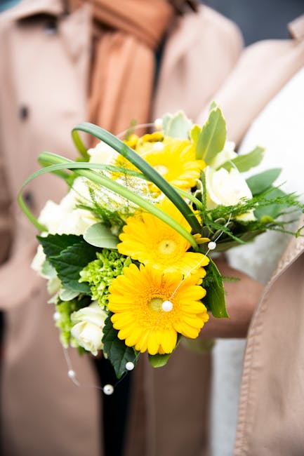 A floral bouquet featuring vibrant yellow gerbera daisies, complemented by white roses and green foliage, held by a person in a beige coat. The arrangement is fresh and lively, with the flowers arrang